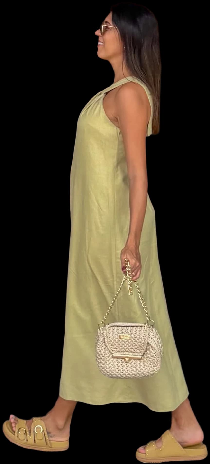 Mulher de vestido verde-claro caminhando em frente a uma porta de madeira entalhada, com bolsa de crochê cru e corrente dourada