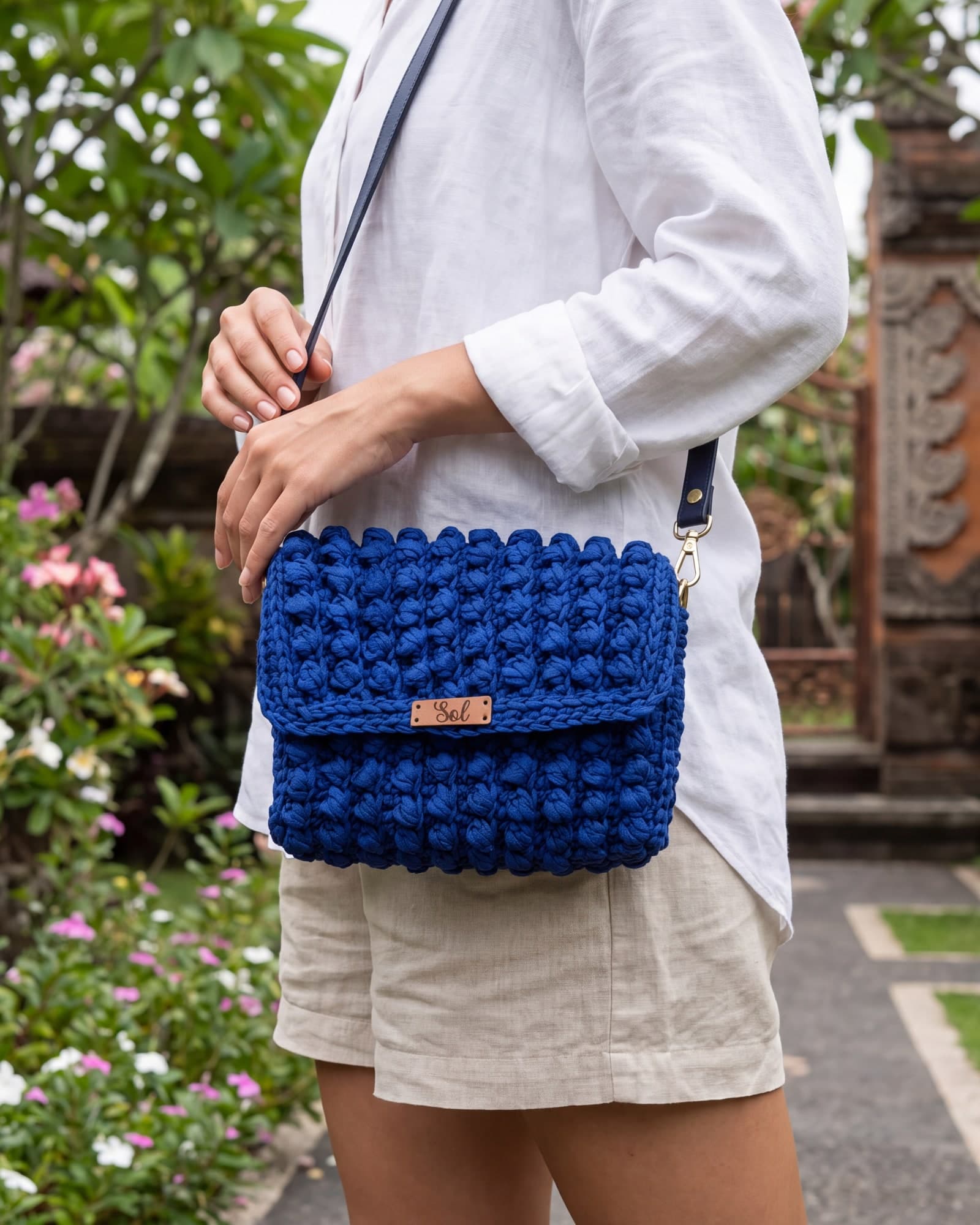 Mulher de camisa de linho branca com a bolsa Bali azul-royal a tiracolo, num jardim com um portal de pedra esculpida, imagem ilustrativa