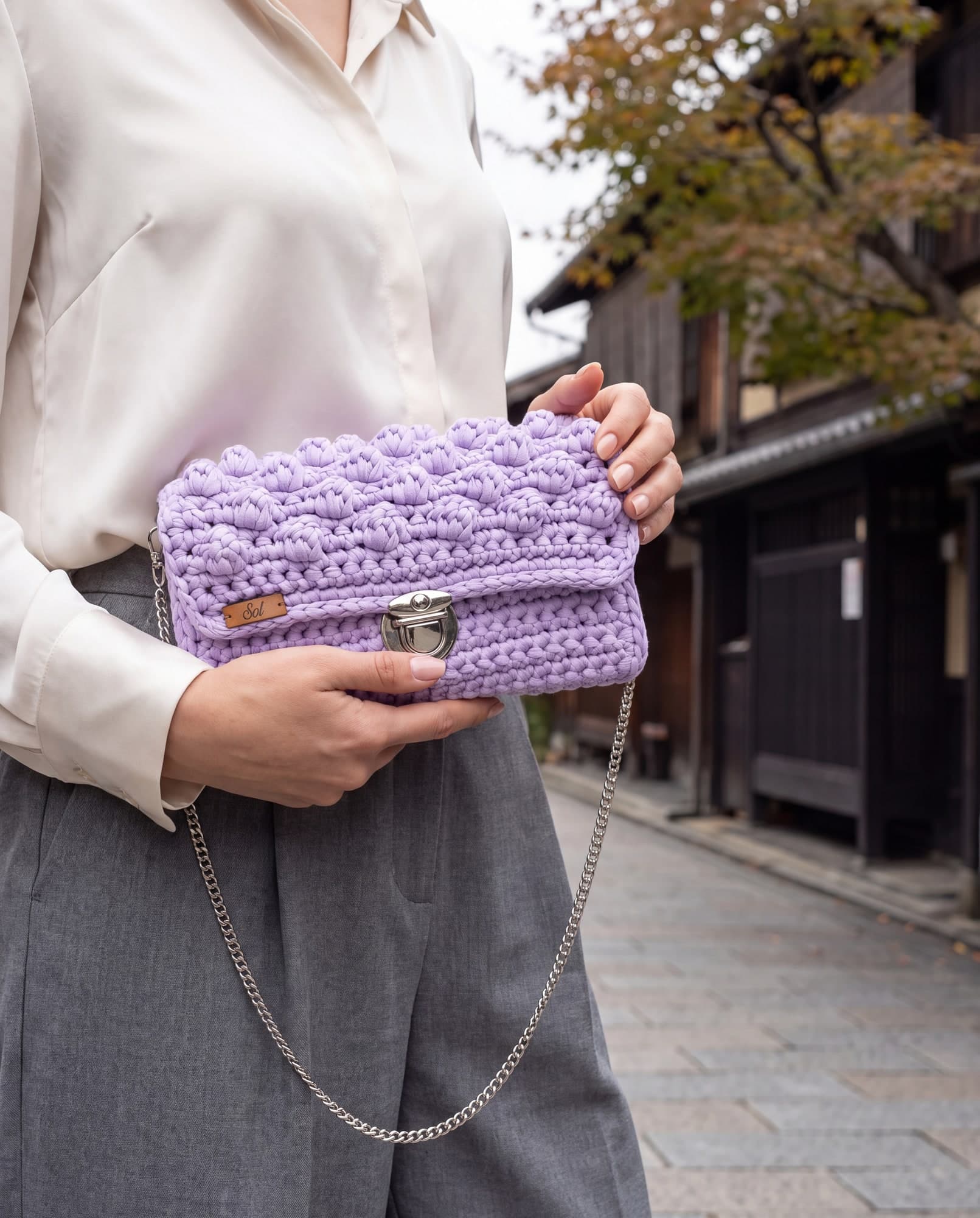 Mulher de blusa marfim segurando a bolsa Kyoto lilás como clutch, numa rua de casas de madeira escura, imagem ilustrativa