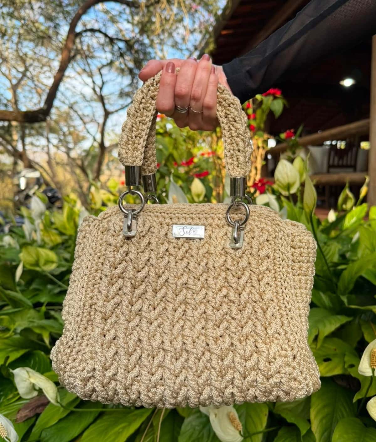 Bolsa Dublin bege em ponto trança, segurada pela alça em um jardim