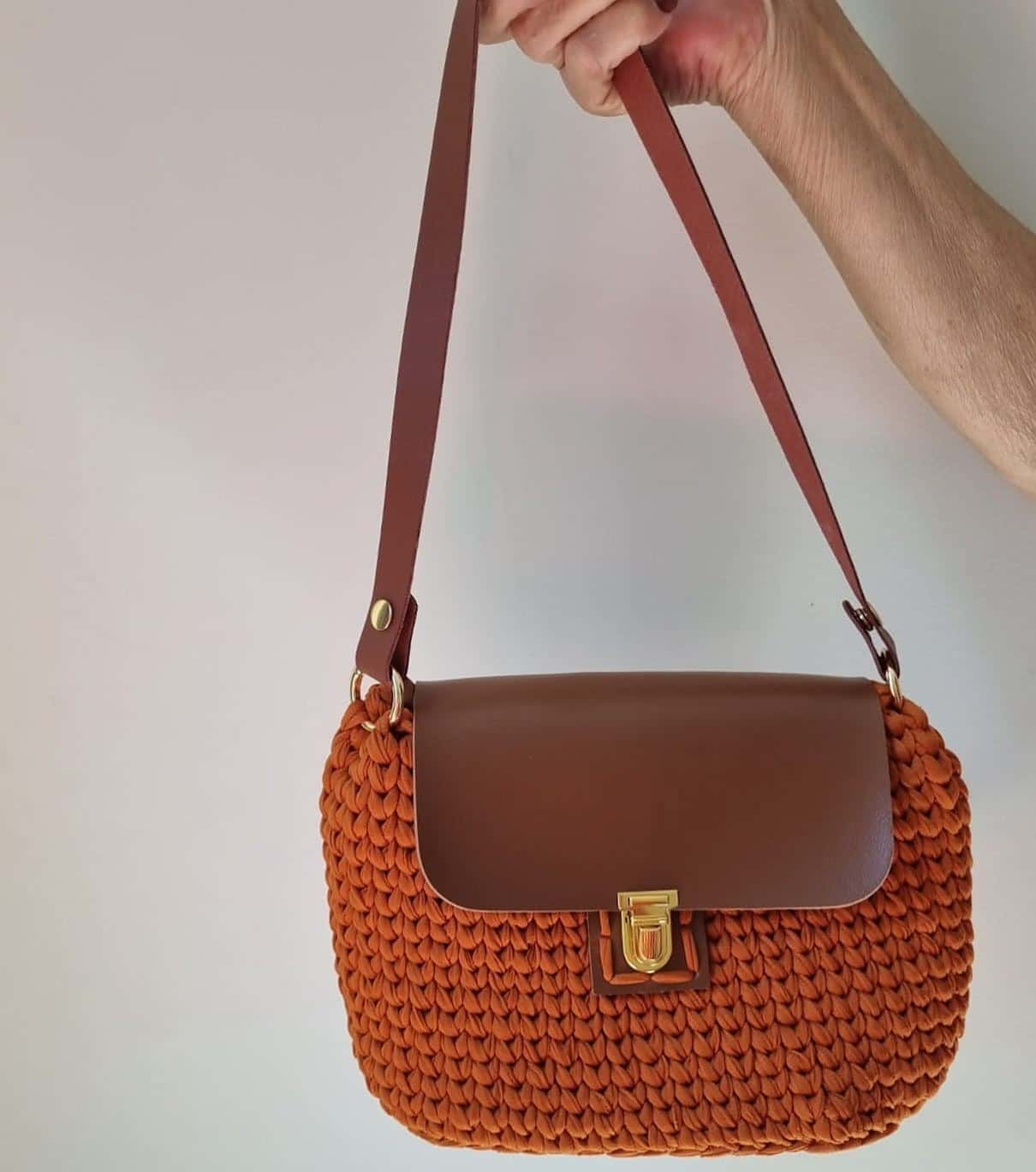 Bolsa Gramado terracota com aba e alça tipo couro caramelo, segurada pela mão