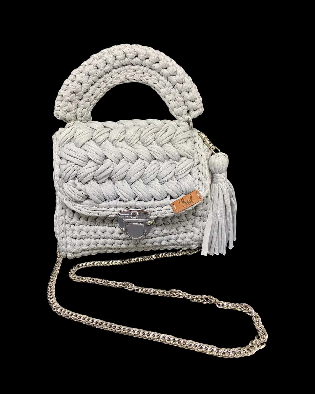 Bolsa Las Vegas cinza com aba trançada, alça de mão em crochê, tassel e corrente prateada, foto de catálogo em fundo neutro