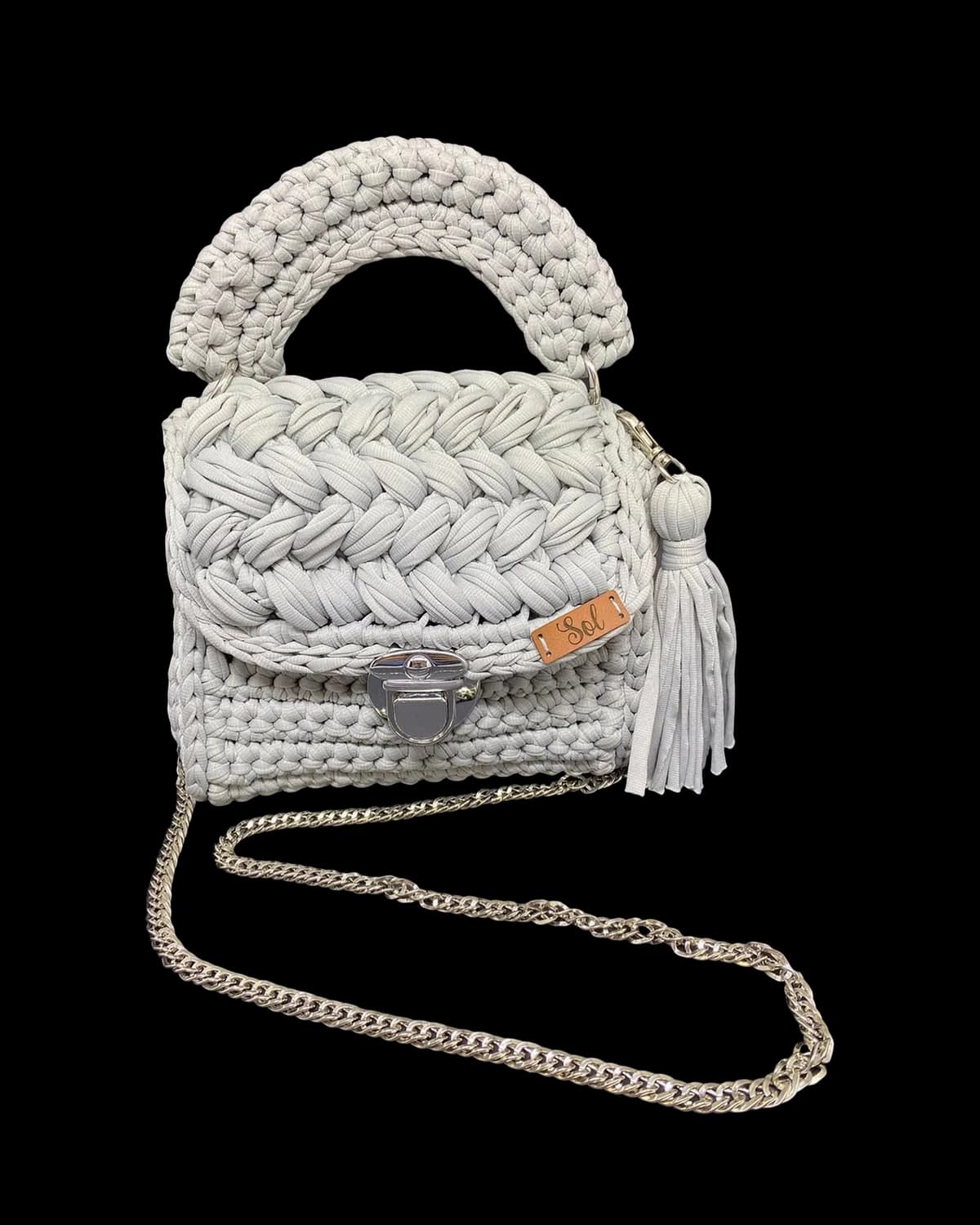 Bolsa Las Vegas cinza com aba trançada, alça de mão em crochê, tassel e corrente prateada, foto de catálogo em fundo neutro